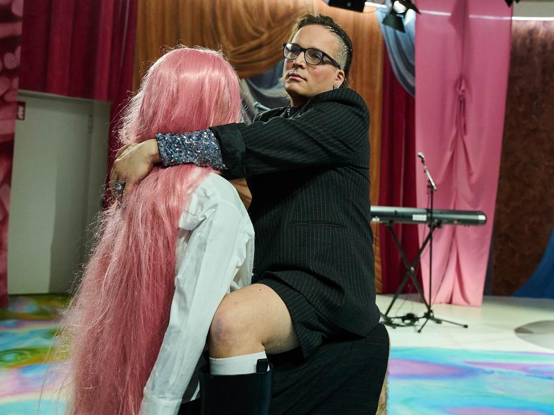 Bühnensituation: Micha Krause sitzt auf dem Schoß einer Puppe. Micha trägt einen schwarzen Anzug mit weißen dünnen Streifen, schwarze Stiefel und weiße Socken; das nackte Knie schaut heraus. Micha Krause trägt eine schwarze Brille und hat die Haare an der Seite wegrasiert und hier mit Glitzer bedeckt. Ihre Arme liegen auf den Schultern der Puppe, die ein weißes Hemd und lange pinke Haare trägt. Im Hintergrund sieht man Stoffe in blau, pink, braun sowie ein Keyboard mit Mikro. 