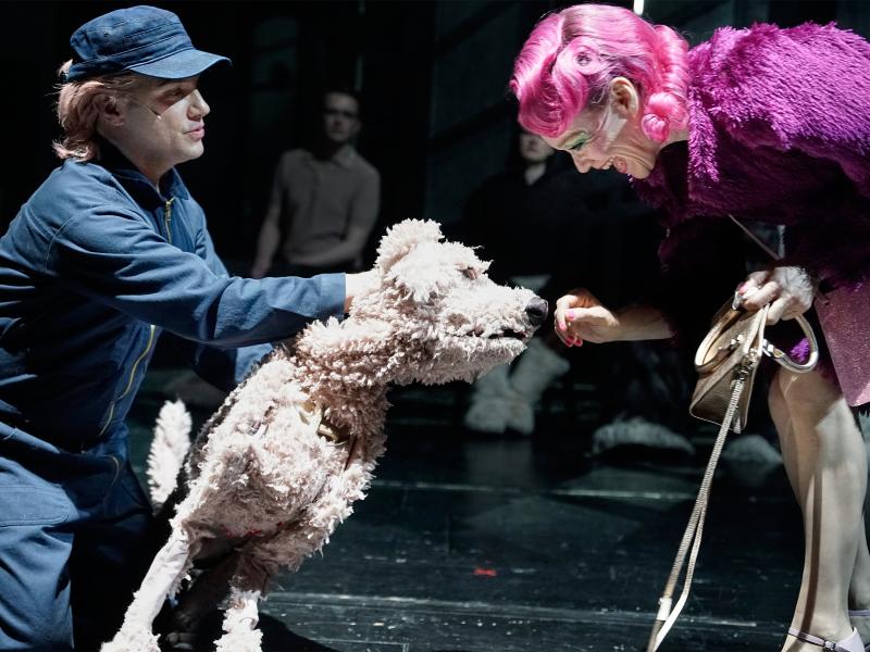 Oscar Olivo im blauen Overall und blauer Schirmmütze und hellen Haaren darunter hat seine Hand an einer lebensgroßen Hundepuppe mit hellem Fell. Der Hund hat seine Schnauze an der Hand von Angelika Richter, die rechts im Bild zu sehen ist und sich zum Hund herunterbeugt. Sie trägt eine pinke Perücke mit Locken, dazu einen lila Fellmantel und eine bronzefarbene Handtasche. Sie lächelt breit. 