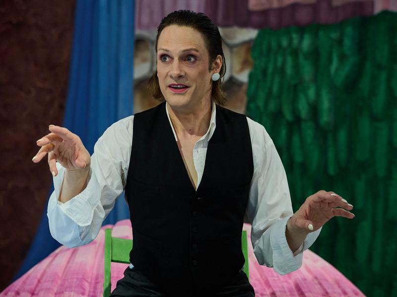 Less Brandes-Hell trägt ein weißes Hemd, darüber eine schwarze Weste, ihre Fingernägel sind weiß lackiert. Sie trägt pinken Lippenstift, große weiße Ohrringe und ihre Augen sind in zartem lila geschminkt und sind weit aufgerissen. Die Arme sind etwas nach vorgestreckt, sie gestikuliert, ihr Mund ist leicht geöffnet. Im Hintergrund sieht man verschiedene bunte Stoffe in pink, blau, grün, lila. 