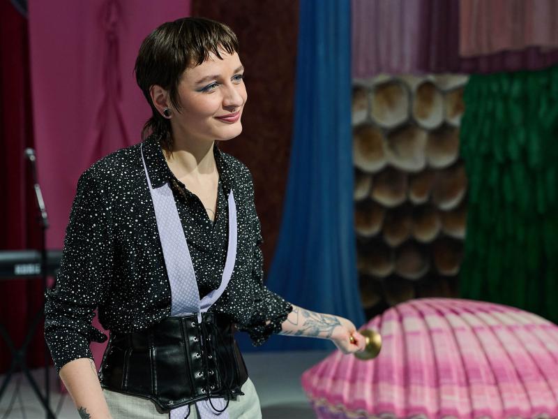 Bühnensituation: Juno Peter trägt ein gepunktetes Hemd und ein Hüftkorsett in schwarzem Leder. Auf Juno Peters Unterarmen erkennt man verschiedene Tattoos. They lächelt und schaut nach rechts. In der einen Hand trägt they einen goldenen Gegenstand. Im Hintergrund erkennt man eine rosafarbene Muschel, sowie verschiedene bunte Stoffe in blau, grün, pink und braun.