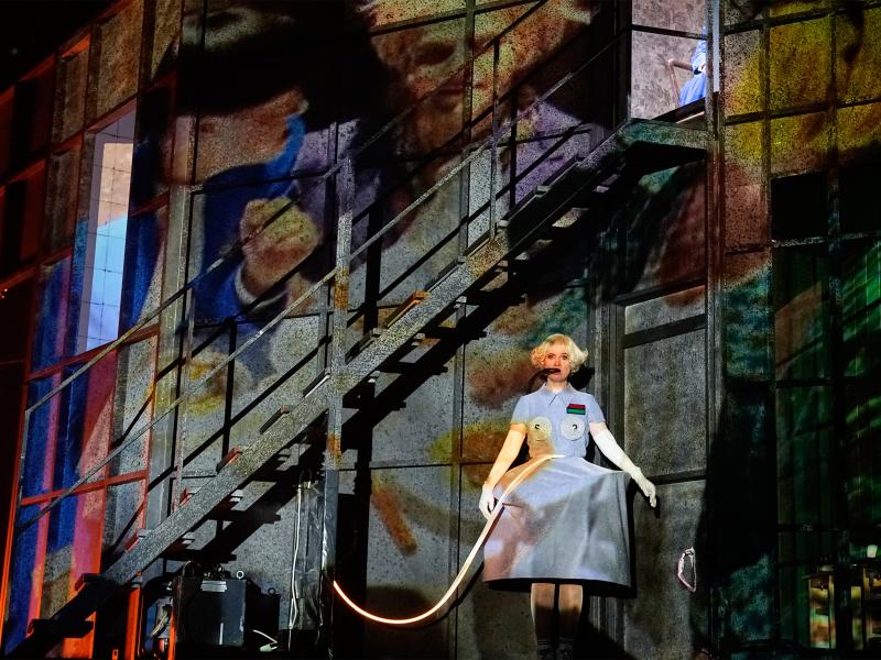 Sandra Gerling ist in der Mitte der Bühne zu sehen, sie hat kurze blonde Locken, trägt ein graues Kleid mit einem runden Reifrock und spitzen Kappen vor den Brüsten und hat einen schwarzen Schlauch im Mund. Ein Kabel ragt auf der Höhe ihres Bauchs in ihr Kleid. Der Rest der Bühne ist dunkel. Man kann eine schwarze Treppe oberhalb von Sandra Gerling erkennen und eine Art Wand aus Kacheln. An die Wand wird ein Video projiziert, auf dem schemenhaft Gesichter zu erkennen sind.