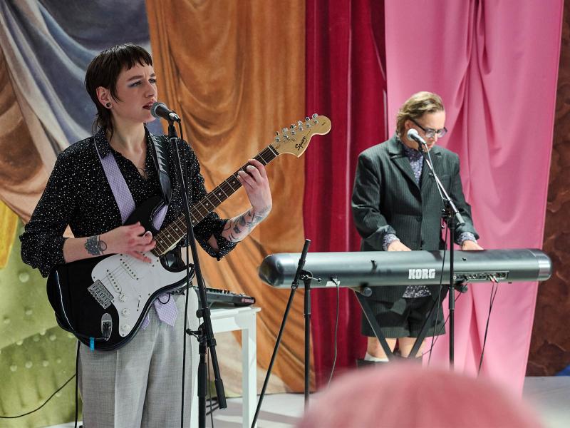 Bühnensituation: Juno Peter hat eine E-Gitarre in der Hand und steht an einem Mikrofon. Der Mund ist leicht geöffnet, die Stirn krausgezogen, der Blick geht in die Ferne. They trägt ein schwarzes Hemd mit weißen Punkten und eine graue Bundfaltenhose. Micha Krause steht hinter einem Keyboard mit Mikro. Sie trägt einen schwarzen Anzug mit kurzer Hose und einem blauen Oberteil darunter. Im Hintergrund sind bunte Stoffe in pink, rot, braun und blau zu sehen.