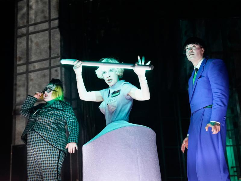 Vor einem schwarzen Hintergrund stehen drei Personen, alle sehen nach vorn. Bettina Stucky trägt einen grün-schwarz karierten Hosenanzug, eine blonde Perücke und eine große Brille mit schwarzem Rahmen. Sandra Gerling steht mittig, hat einen kurzen blonden Bob, trägt ein graues Kleid mit einem Reifrock und spitzen Kappen vor den Brüsten. In den Händen hält sie eine Neonröhre. Rechts steht Maximilian David Scheidt in blauem Anzug, seine Haare sind schwarz. Alle Münder sind leicht geöffnet.