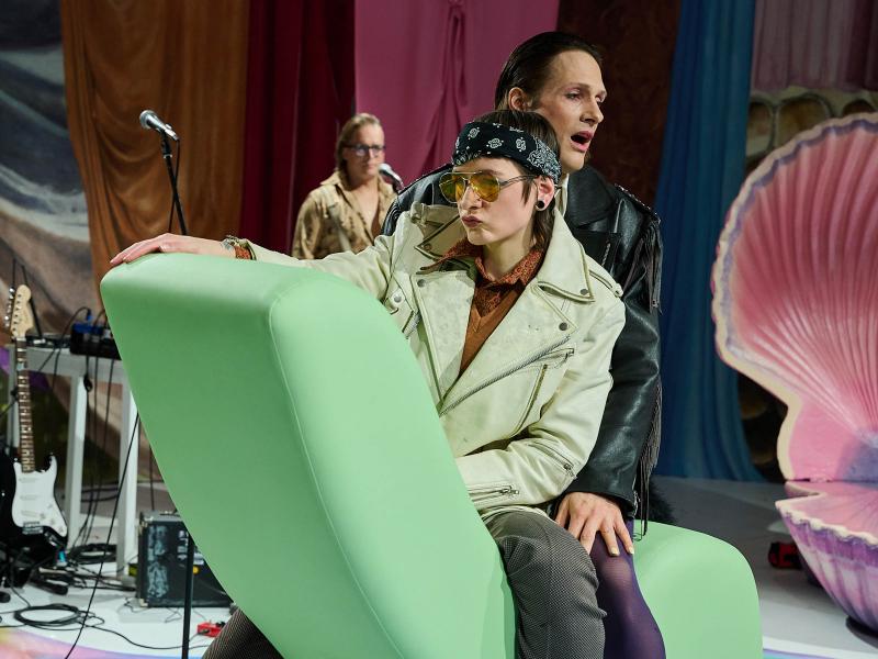 Bühnensituation: Juno Peter und Less Brandes-Hell sitzen hintereinander auf einem grünen Sofa, dessen lange Seite in die Luft gekippt ist. Beide tragen Lederjacken mit Fransen, Juno sitzt vorn und trägt ein Bandana-Tuch um die Haare und eine Fliegersonnenbrille. Junos Lippen sind gespitzt, their rechte Hand liegt auf der Sofaspitze. Im Hintergrund steht Micha Krause an einem Mikrofon, eine E-Gitarre ist zu sehen und eine große aufgeklappte rosa Muschel.