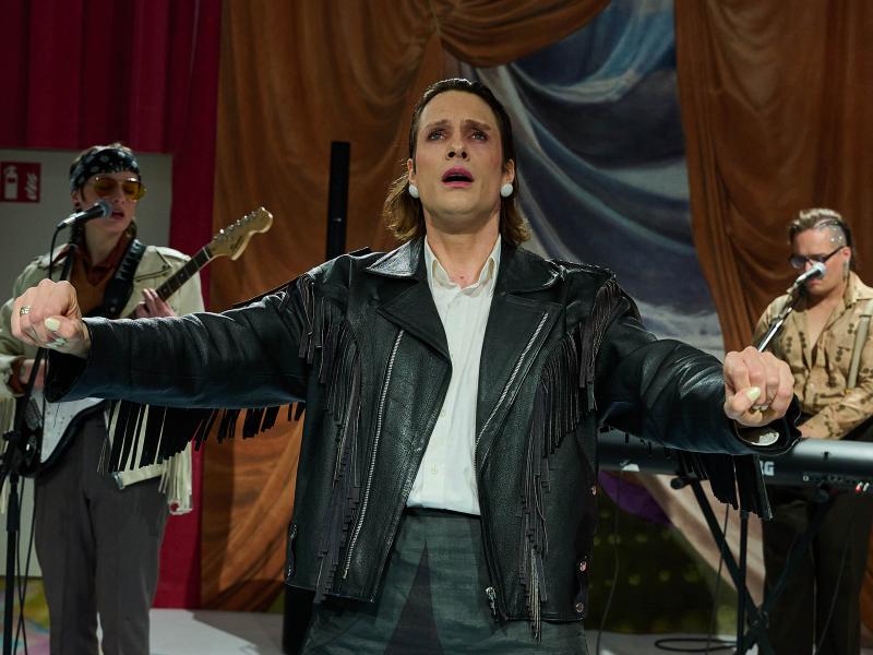 Bühnensituation: Less Brandes-Hell trägt eine schwarze Fransen-Lederjacke, ihre Arme sind nach vorne ausgestreckt, als würden sie etwas umfassen. Ihr Mund ist geöffnet, sie schaut nach vorn. Im Hintergrund stehen Juno Peter mit E-Gitarre und Micha Krause am Keyboard, Juno singt in ein Mikrofon und trägt ebenfalls eine Lederjacke.