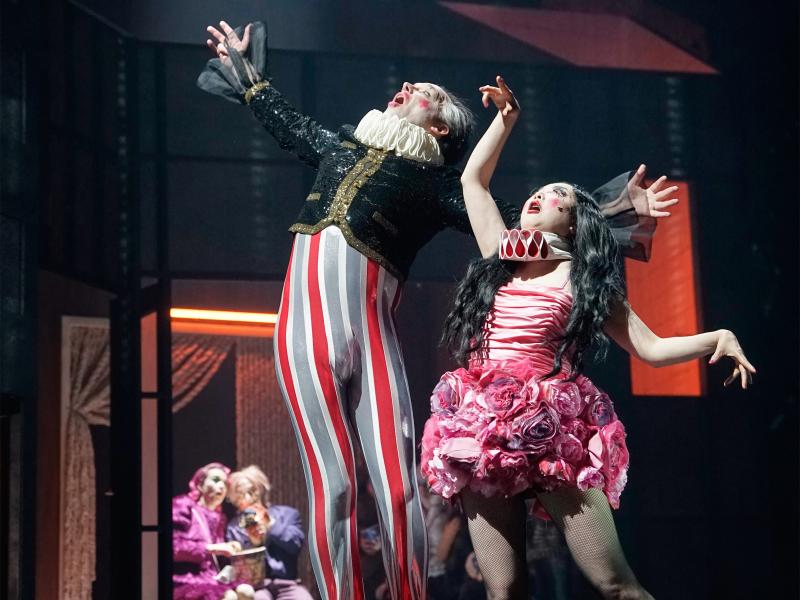 Im Vordergrund sind Felix Knopp und Sachiko Hara zu sehen. Er trägt eine grau-weiß-rote enge Hose und rote Clownsschuhe, eine schwarze Weste mit Pailletten und eine weiße Krause um den Hals. Augen und Mund sind weit aufgerissen, er schaut nach oben und hat beide Arme seitlich ausgestreckt. Sachiko Hara trägt ein Kleid, das mit Rosen besetzt ist. Ihre Krause ist rosa, ihre langen schwarzen Haare sind offen. Sie schaut hoch, Mund und Augen weit auf und die Arme sind nach oben und zur Seite gestreckt. 