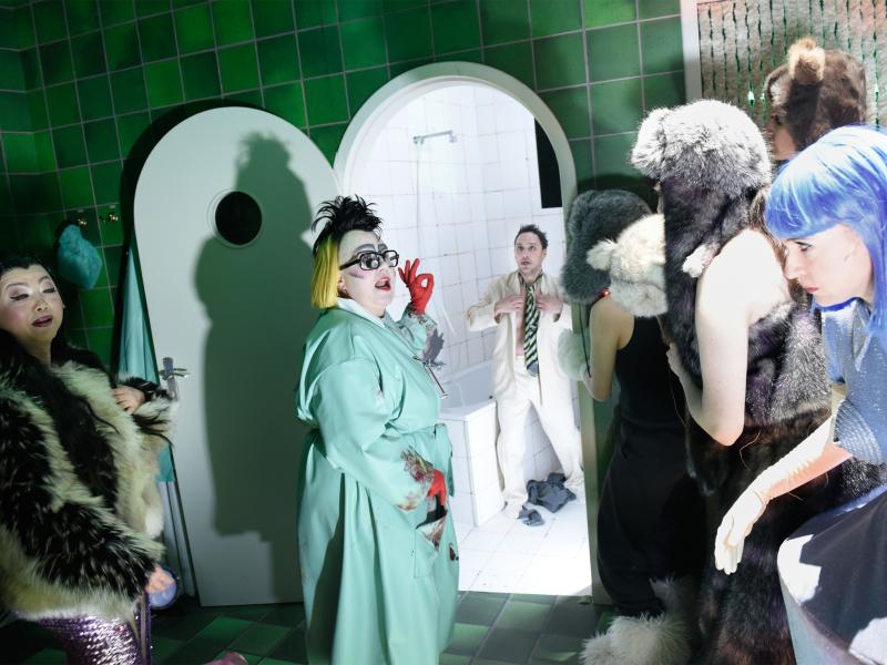 Im Zentrum steht Bettina Stucky in einem türkisen Kittel und roten Handschuhen, ihre Haare sind schwarz und blond. Ihre große Brille hat einen schwarzen Rahmen, ihr Mund ist geöffnet. Sie dreht sich zu Personen in Fellkleidung und Fellmütze. Vor ihnen steht Sandra Gerling in blauer Perücke und blau-grauem Kleid sehen. Links im Bild steht Sachiko Hara in einer Felljacke und mit geschlossenen Augen. Im Hintergrund sieht man Oscar Olivo in einem Kachelraum mit Badewanne, er trägt einen weißen Anzug.