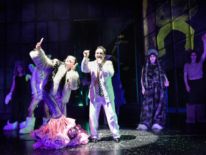 Im Vordergrund stehen Sachiko Hara und Oscar Olivo. Sie trägt eine Felljacke und darunter einen Rock aus lila Pailletten mit Tüll ab den Knien, sodass die Füße verdeckt sind. Sie hält Zeige- und Mittelfinger hoch und macht ein Selfie mit einem Handy. Sie lächelt. Oscar Olivo trägt einen weißen Anzug und eine schwarz-gelb gestreifte Krawatte. Er hat eine Hand in die Höhe gestreckt, mit der anderen hält er ein Mikrofon an seinen geöffneten Mund. Im dunklen hinteren Bühnenbereich sieht man die Chormitglieder. 