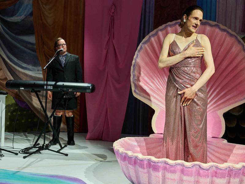 Bühnensituation: Less Brandes-Hell steht in einer rosafarbenen aufgeklappten Muschel. Sie trägt ein rosa Glitzerkleid, hält sich eine Hand vor die Brust und eine Hand vor den Schritt. Sie hat den Mund leicht geöffnet und schaut nach rechts. Im Hintergrund steht Micha Krause hinter einem Keyboard und am Mikro. Sie trägt einen schwarzen Anzug mit einem blauen Oberteil darunter sowie schwarze Stiefel. Der Hintergrund ist mit bunten Stoffen verhangen.