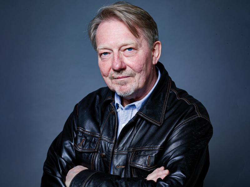 Dietmar Wischmeyer steht vor einem grau-blauen Hintergrund. Er trägt kurze grau-braune Haare, ein hellblaues Hemd und eine schwarze Lederjacke. Er hat die Arme vor der Brust verschränkt und schaut in die Kamera.