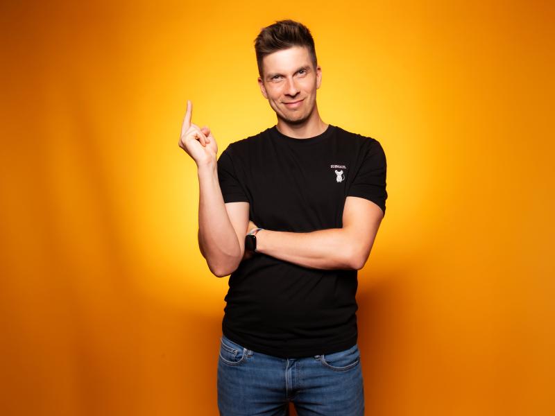 Simon Stäblein steht vor einem orangen Hintergrund. Er trägt kurze braune Haare und ein schwarzes T-Shirt. Er hat den linken Arm vor der Brust verschränkt, den rechten Arm hat er angewinkelt und zeigt mit dem Zeigefinger derselben Hand in die Luft. Er schaut in die Kamera und lächelt.