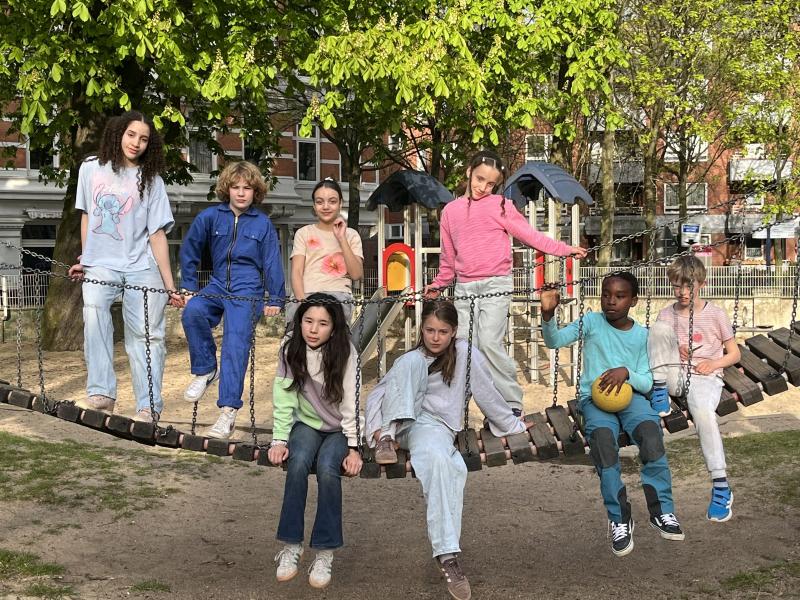 Zu sehen sind acht Kinder, die auf einer Hängebrücke eines Spielplatzes stehen und sitzen. Sie tragen unterschiedliche, bunte Kleidung und schauen in die Kamera. Im Hintergrund sind Bäume zu erkennen.