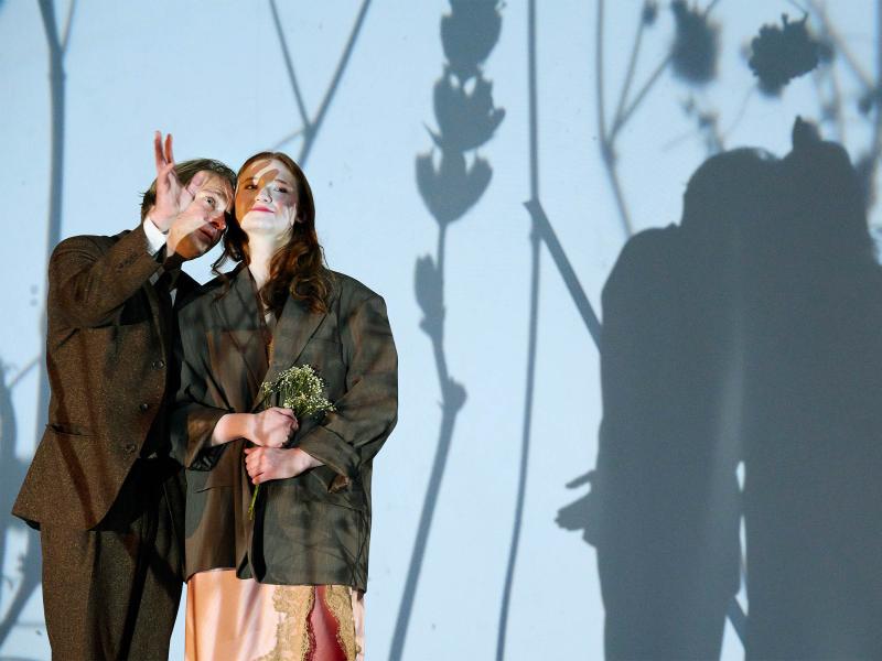 Bühnensituation: Emma Oberpichler trägt ein rosafarbenes Kleid und ein großes braunes Jackett darüber. Sie hält einen kleinen Strauß Blumen in der Hand. Dicht neben ihr steht Mirco Kreibich in einem braunen Anzug. Er hält sein Gesicht nah an ihre Wange und hebt die Hand, um zu gestikulieren. Hinter ihnen ist eine große weiße Wand, auf denen die Schatten der beiden zu sehen sind.