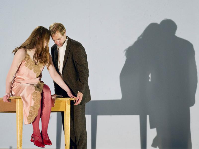 Bühnensituation: Emma Oberpichler trägt ein rosafarbenes Kleid mit goldfarbenen Verzierungen. Sie hat die Haare offen und sitzt auf einem großen Holztisch. Neben ihr am Tisch steht Mirco Kreibich und beugt sich zu ihr. Ihre Hände berühren sich und sie haben die Köpfe nah beieinander. Hinter ihnen ist eine große weiße Wand. Dort sind die Schatten der beiden und des Tisches. 