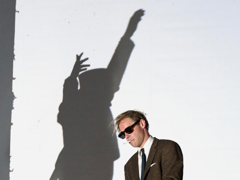 Bühnensituation: Mirco Kreibich mit braunem Anzug mit grüner Krawatte, Sonnenbrille und Zigarette im Mund, seine Arme und ein Bein sind leicht angewinkelt. Im Hintergrund der Schatten einer Person im kniefreien Kleid, die beide Arme erhoben hat, auf einer weißen Wand.