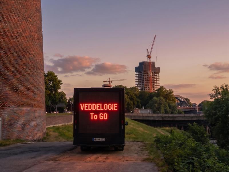Außenansicht. Links im Bild ist ein großes rotes Backsteingebäude zu erkennen. Davor, auf einem rauen Betonuntergrund, parkt ein schwarzer LKW mit einer großen Leuchtschrift auf der Rückseite. Dort steht: VDDLTOPOLY to GO. Im Hintergrund ist eine Flussumgebung des Hamburger Hafens mit einer Brücke und Autos zu sehen. Ganz im Hintergrund ragt der unfertige bau des Elbtowers mit zwei großen Kränen in den Himmel.