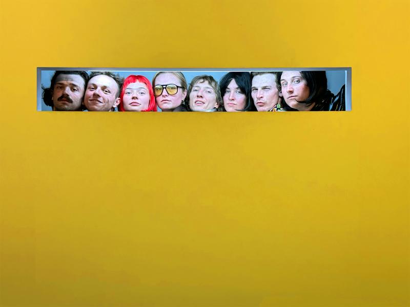 Eine gelbe Wand: In einem kleinen rechteckigen und sehr langgezogenem Fenster sind acht Menschen zu sehen. Man sieht von ihnen nur die Gesichter. Sie schauen in die Kamera.