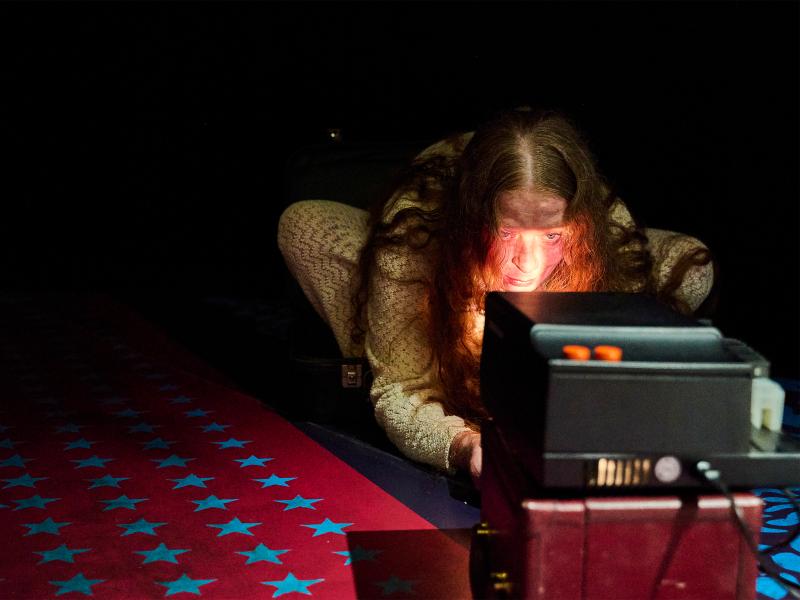 Christiane von Poelnitz hockt vor einem Projektor und schaut in das Licht, das aus ihm scheint. Sie trägt ein langärmliches, beiges Strickkleid, ihre offenen Haare fallen ihr über Schulter und Arme. Der Boden ist rot und hat Reihen aus blauen Sternen darauf. Der Hintergrund ist dunkel.