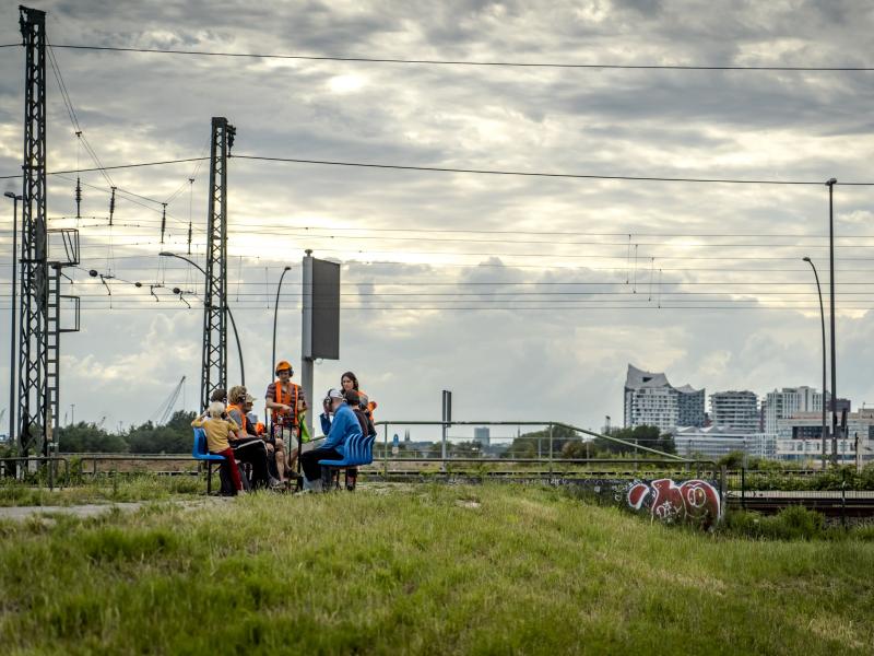 Zu sehen ist eine Menschengruppe, die auf einer blauen Sitzbank auf einer Wiese sitzt. Einige Personen stehen. Sie tragen alle Kopfhörer. Zwei Personen tragen eine orangene Warnuniform. Im Hintergrund sind Bahngleise und die Elbphilharmonie zu sehen.