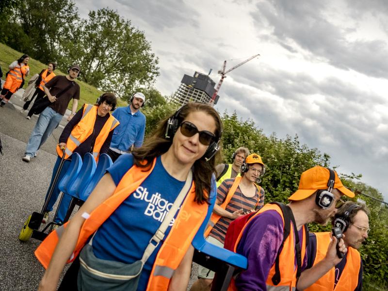 Mehrere Personen in orangener Warnuniform tragen eine blaue Sitzbank, sie tagen alle Kopfhörer und laufen dabei eine Straße entlang. 