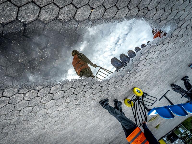 Eine Person mit orangene Warnuniform schiebt eine Sackkarre vor sich, die Spiegelung der Person ist in einer Pfütze zu sehen. 