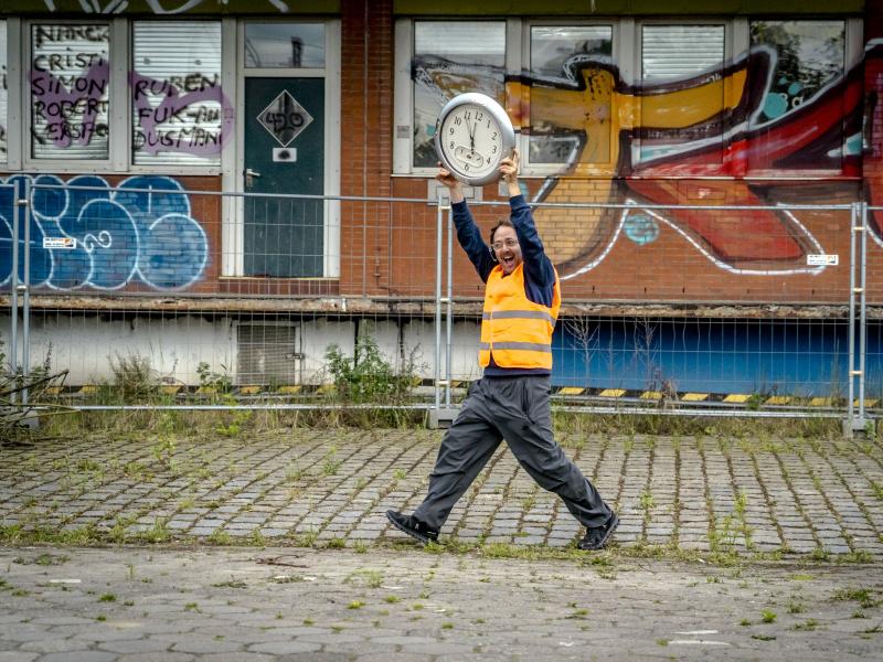 Samuel Weiss läuft in orangener Warnuniform an Baustellenzäuen entlang, dahinter eine mit Graffiti bemaltes Gebäude. Samuel Weiss hält eine große Uhr hoch über seinen Kopf.