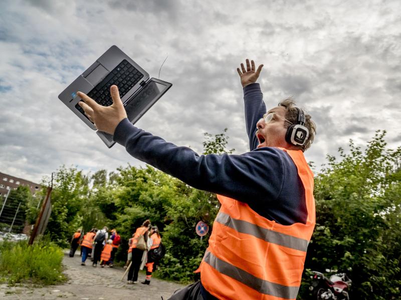 Samuel Weiss läuft in orangener Warnuniform eine Straße entlang vor ihm sind mehrere Personen in orangener Warnuniform. Ein Laptop ist gerade dabei auf den Boden zu fallen. Samuel Weiss schaut erschrocken.