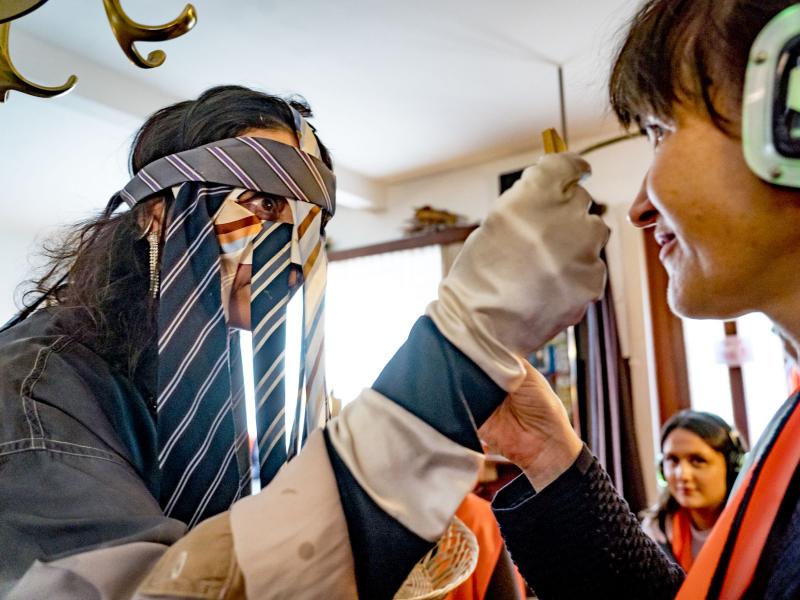 Sasha Rau trägt eine Maske aus Krawatten, sie hält etwas vor die Augen einer Person, die eine orangene Warnuniform und Kopfhörer trägt. 