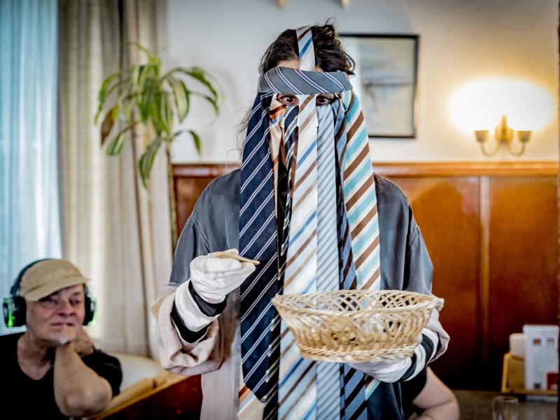 Sasha Rau trägt eine Maske aus Krawatten, sie hält einen Korb und ein Stück Holz in den Händen, im Hintergrund sitzt ein Mann mit Kopfhörern auf dem Kopf. 