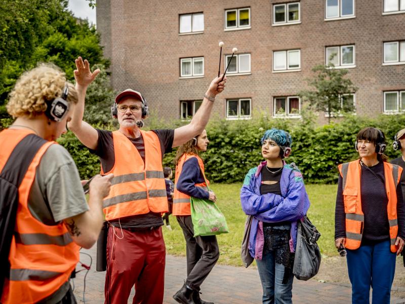 Mehrere Personen in orangener Warnuniform und Kopfhörern stehen verteilt in einem Wohngebiet. Yorck Dippe steht mit ausgestreckten Armen da.