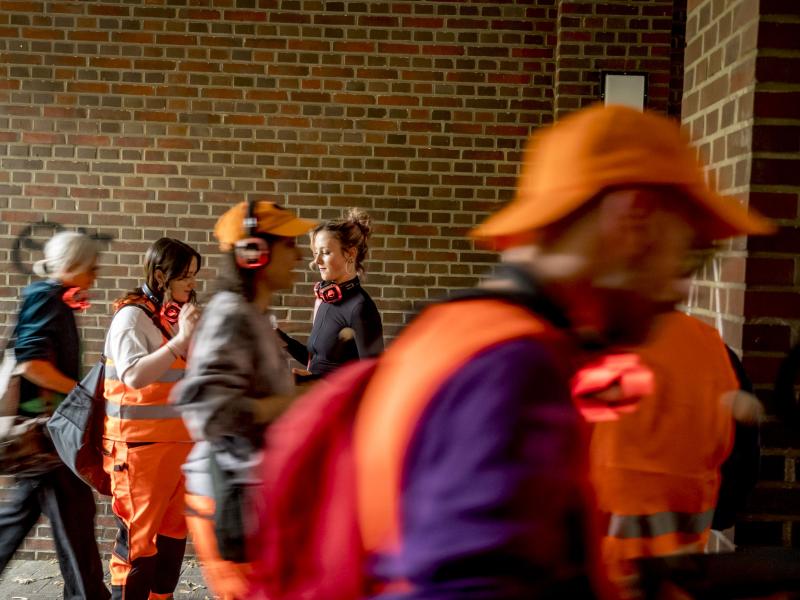 Mehrere Personen in orangener Warnuniform und Kopfhörern laufen durch eine überdachte Unterführung.
