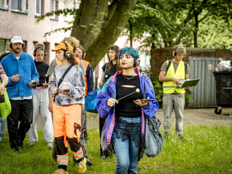 Mehrere Personen mit Kopfhörern laufen über eine Wiese, mache von ihnen tragen orangene Warnuniformen und haben Schläger in der Hand.