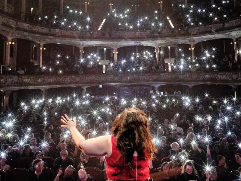 Eine Person mit langen braunen Haaren und rotem Oberteil steht von hinten fotografiert auf einer Bühne und hebt den rechten Arm zum Publikum. Im vollbesetzten, prunkvollen Theatersaal mit mehreren Rängen halten hunderte Zuschauer ihre beleuchteten Smartphone-Taschenlampen in die Höhe, die im Bild als funkelnde Lichtsterne erscheinen.