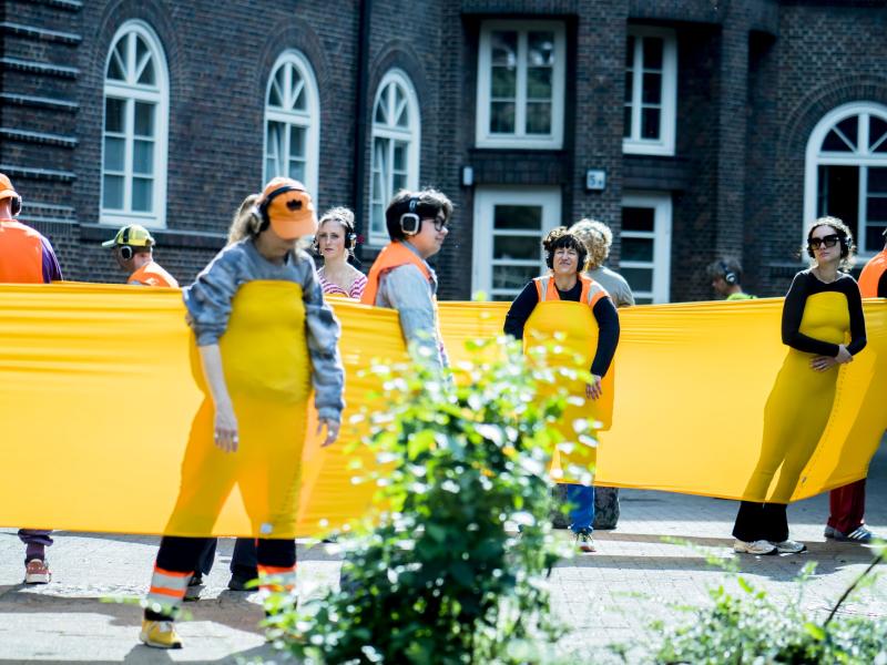 Mehrere Personen mit Kopfhörern befinden sich in einem überdimensional großen gelben Gummiband. Alle Personen lehnen sich nach außen an das Gummiband, sodass es unter Spannung steht. Sie befinden sich vor einem Wohngebäude. 