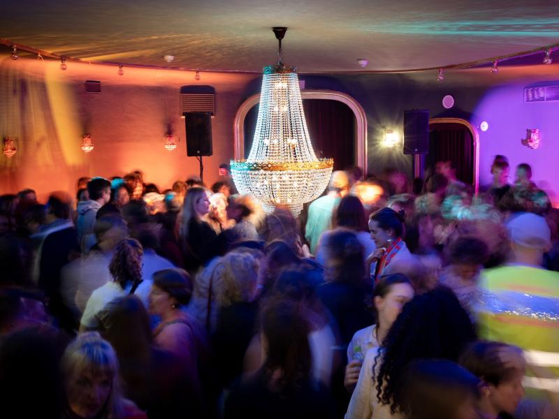 Ein dicht gefüllter Veranstaltungsraum mit vielen Menschen, die sich bewegen und miteinander sprechen. In der Mitte hängt ein großer, beleuchteter Kristalllüster von der Decke. Die farbige Beleuchtung in Orange-, Rosa- und Blautönen erzeugt eine stimmungsvolle Atmosphäre. Durch die lange Belichtungszeit wirken viele Personen verschwommen, wodurch die Dynamik und Bewegung der Menschenmenge betont wird.