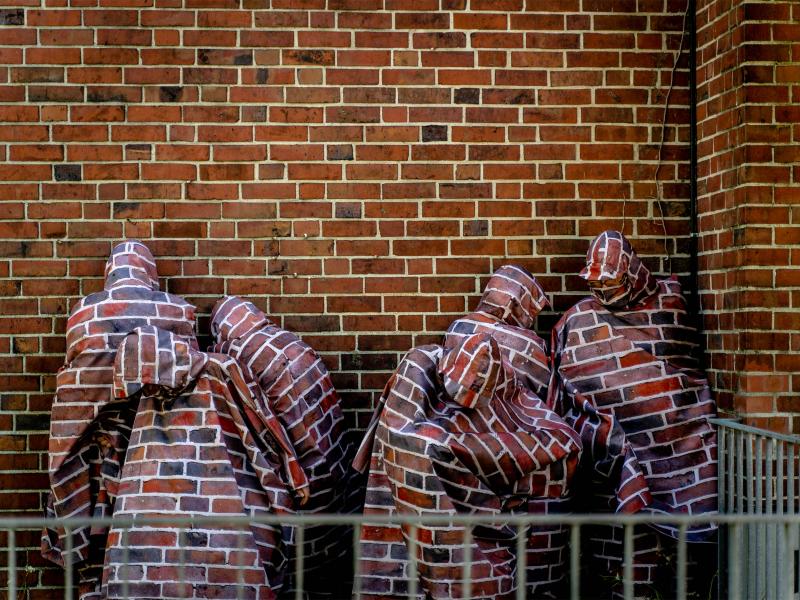 Vor einer Backsteinmauer stehen vier Körper. Sie tragen Umhänge mit Kapuzen, die mit dem Muster der Backsteinmauer bedruckt sind. Die Gesichter sind nicht zu erkennen.