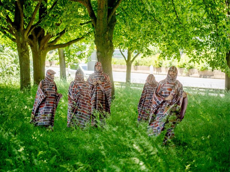 Fünf Menschen stehen auf einer grünen Wiese unter grünen Bäumen. Sie tragen Umhänge mit Kapuze, die mit dem Muster einer Backsteinmauer bedruckt sind.