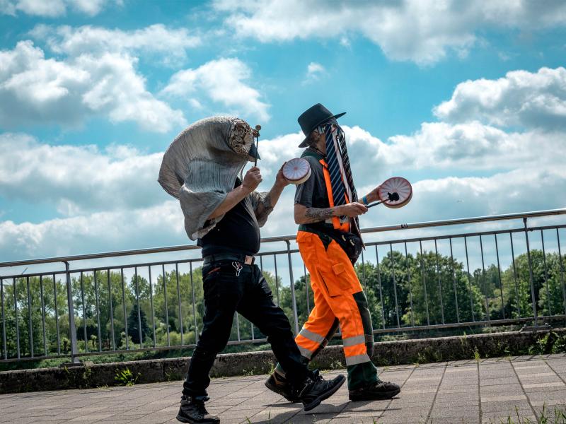 Zu sehen sind zwei Menschen, die auf Asphalt an einem Geländer entgehen. Hinter ihnen sieht man Bäume und Wolkenhimmel. Eine Person hat schwarze Kleidung, eine übergroße vogelähnliche Maske. Die andere Person trägt eine orangene Hose mit reflektierenden Streifen und einen Hut mit Krawatten daran. Beide halten eine kleine Trommel in der Hand.