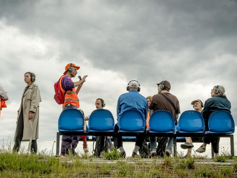 Zu sehen ist eine Menschengruppe, die auf einer blauen Sitzbank auf einer Wiese sitzt. Einige Personen stehen. Sie tragen alle Kopfhörer. Eine Person trägt eine orangene Warnuniform. 