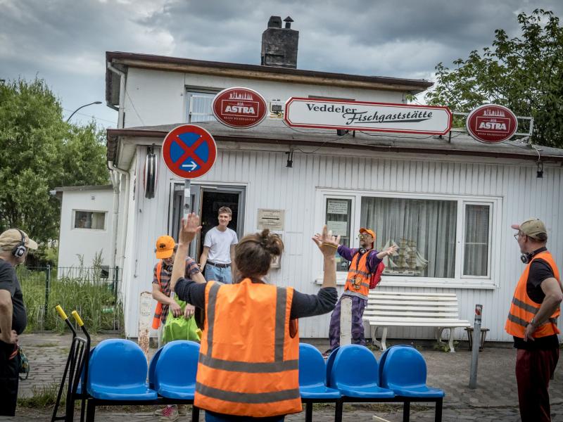 Mehrere Personen in orangener Warnuniform stehen verteilt vor der „Veddeler Fischgaststätte“ mit ausgestreckten Armen.