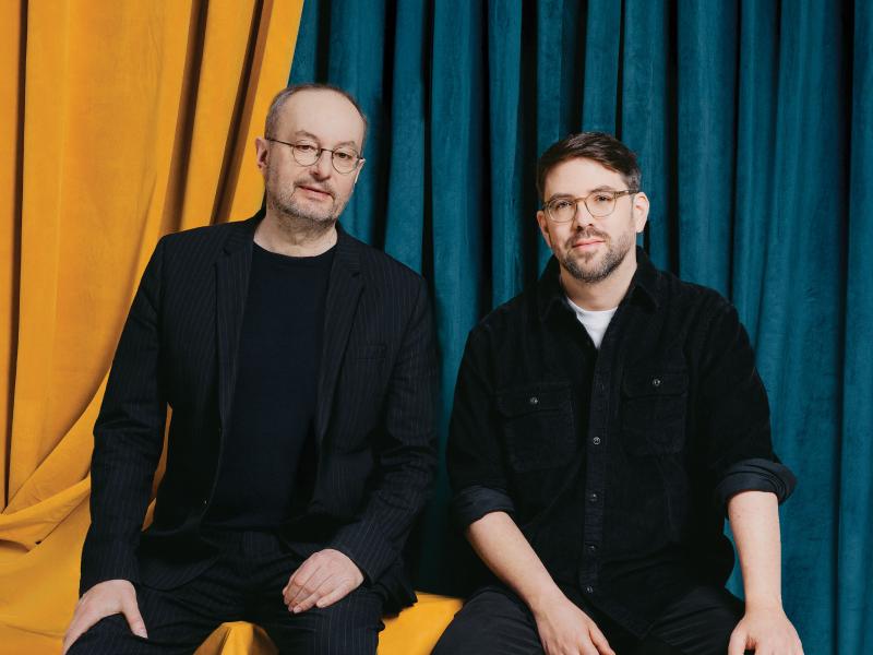 Joachim Telgenbüscher und Nils Minkmar sitzen nebeneinander vor einem Hintergrund aus farbigen Samtvorhängen in Ocker und Petrol. Nils Minkmar trägt ein dunkles Nadelstreifen-Sakko und runde Metallbrille, Joachim Telgenbüscher ein schwarzes Cordhemd und eine markante Hornbrille mit hellem Rahmen. Beide blicken ruhig in die Kamera. Das Foto wirkt wie ein professionelles Porträt, etwa für ein Theater- oder Kulturprogramm.