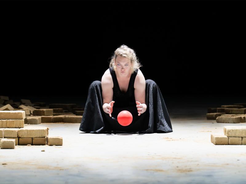 Ankündigungsfoto »Fremde Sonne II: Medea in Korinth«; Lina Beckmann in schwarzem Gewand hockt in der Mitte einer kargen Bühne und beugt sich über eine leuchtend rote Kugel, die Hände wie beschwörend darüber ausgestreckt. Rechts und links gestapelte Ziegelsteine, der Hintergrund vollständig im Dunkel. Die Totale betont die Isolation der Figur im leeren Bühnenraum.
