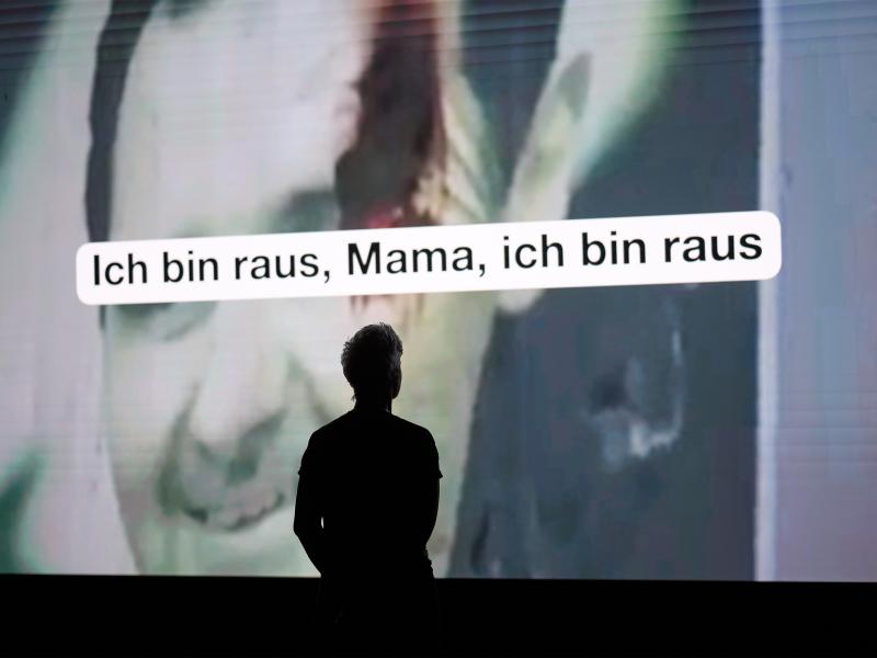 Person als Silhouette vor einer großen Videoprojektion; darüber steht in einem weißen Textfeld: „Ich bin raus, Mama, ich bin raus“.