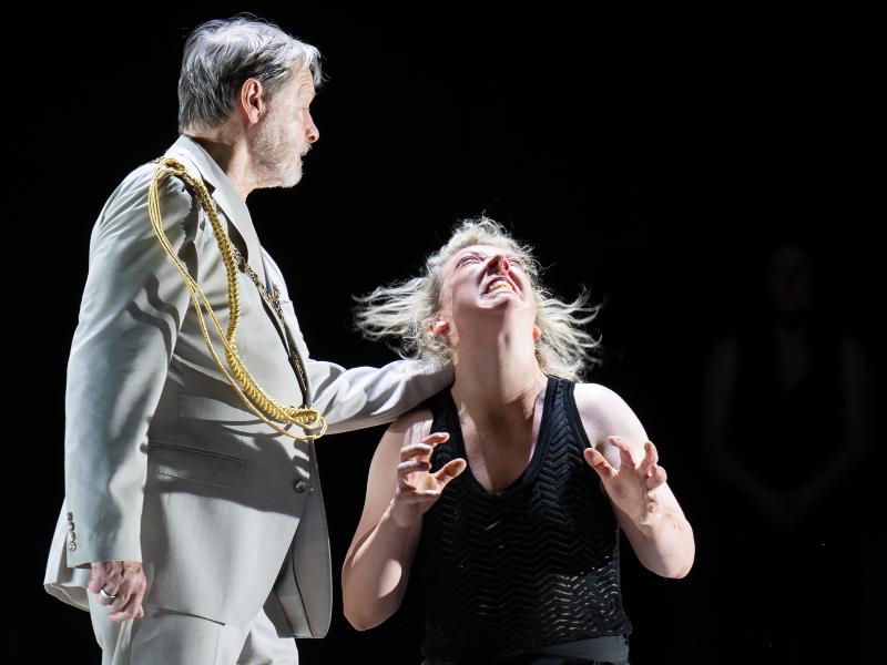 Ankündigungsfoto »Fremde Sonne II: Medea in Korinth«; Lina Beckmann im schwarzen Top reißt den Kopf zurück, die Hände verkrallt – ein Ausdruck extremer Qual oder Raserei. Michael Wittenborn im hellen Anzug mit goldener Schulterschnur greift ihr von hinten an die Schulter.
