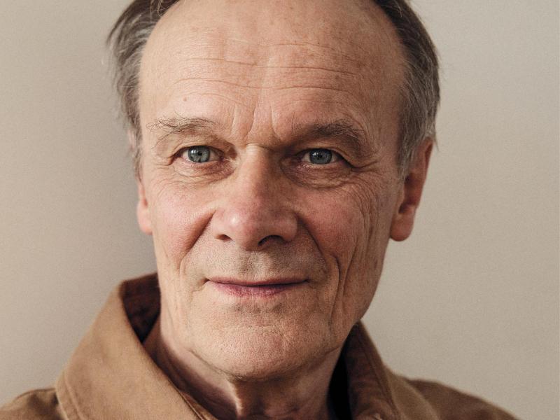 Nahaufnahme von Schauspieler und Autor Edgar Selge. Er trägt kurze graue Haare und ein helles Hemd. Er blickt leicht an der Kamera vorbei und lächelt leicht vor einem neutralen, hellen Hintergrund.