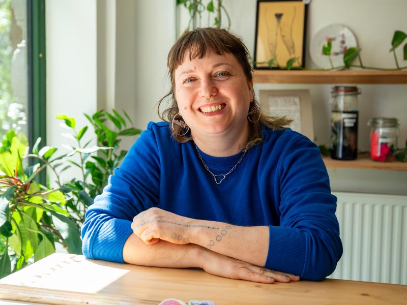 Porträt einer lächelnden Person in blauem Pullover und mit Schmuck, an einem Holztisch in einem hellen, mit Pflanzen eingerichteten Raum.