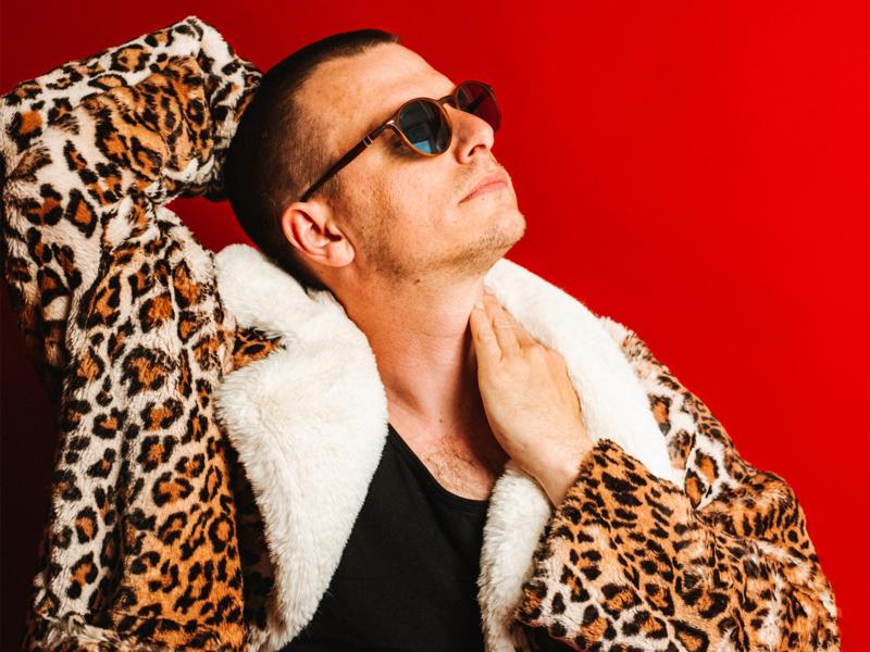 Porträt von Moritz Neumeier mit Sonnenbrille, schwarzem Oberteil und auffälligem Leopardenmantel mit hellem Fellkragen. Die Person posiert vor einem leuchtend roten Hintergrund.