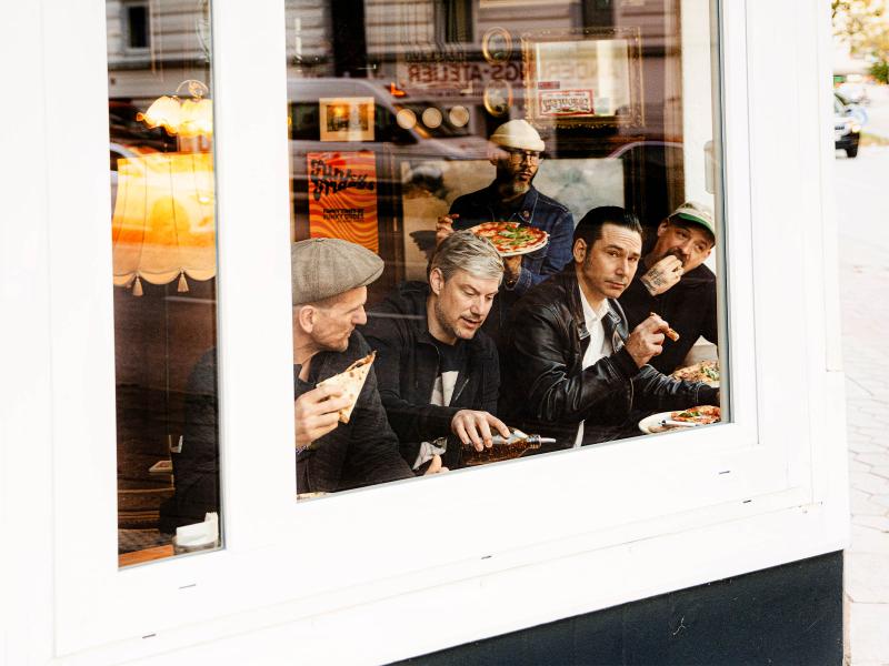 Fünf Männer sitzen in einem Restaurant hinter einer großen Fensterscheibe und essen Pizza. Einer der Männer hält eine Pizza hoch, während die anderen Pizza­stücke in der Hand halten. Die Männer tragen überwiegend dunkle Kleidung und teilweise Schiebermützen oder eine Kappe.