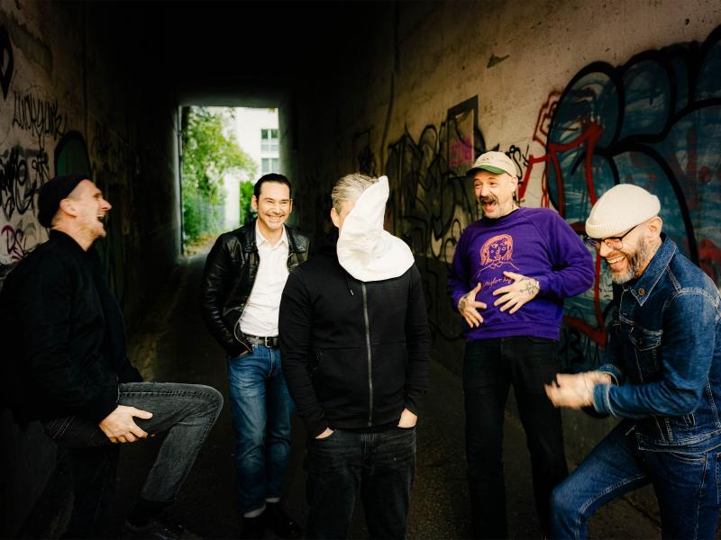 Fünf Männer stehen lachend in einem Tunnel mit Graffiti-Wänden. Eine Person in der Mitte kehrt der Kamera den Rücken zu, die Kapuze hochgezogen. Die anderen vier sind ausgelassen und bewegt, die Stimmung locker und humorvoll.