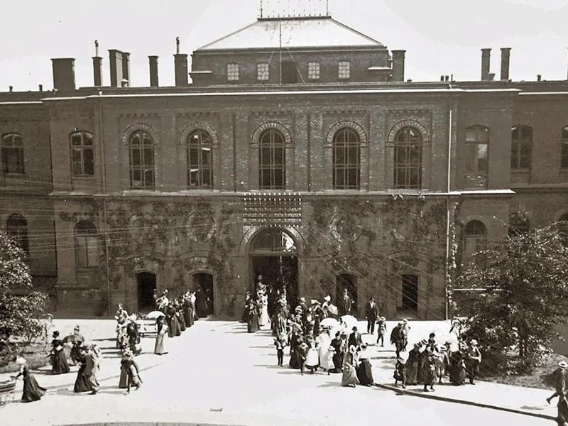 Historische Schwarzweißaufnahme des Universitätsklinikums Hamburg-Eppendorf (UKE). Vor einem repräsentativen Klinikgebäude mit großen Rundbogenfenstern stehen und gehen zahlreiche Menschen, darunter viele Frauen und Kinder. Die Aufnahme zeigt das UKE in historischer Zeit.